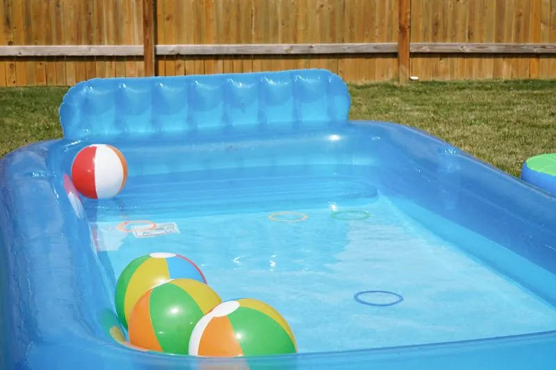 Petite piscine gonflable remplie d'eau, avec des ballons de plage, sur la pelouse d'un jardin clôturé