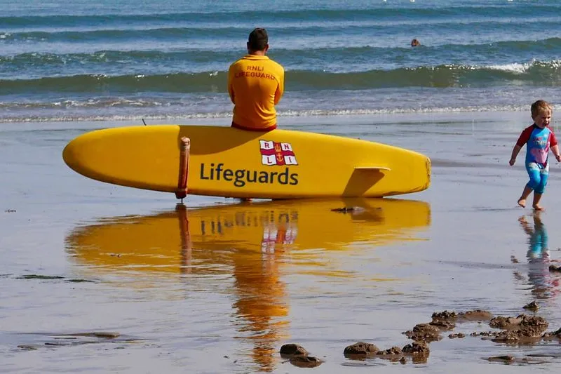 Sauveteur assis derrière une planche de sauvetage jaune, face à la mer, pendant qu'un jeune enfant court sur le sable mouillé