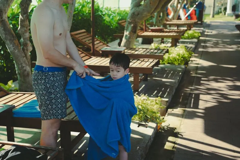 Un père enveloppe son jeune fils dans une grande serviette bleue, à côté des transats d'une piscine