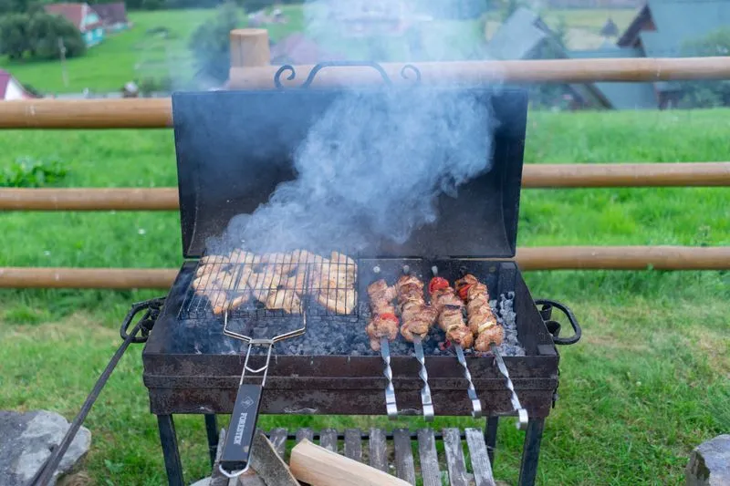 Barbecue rectangulaire à charbon, couvercle relevé, morceaux de poulet dans une grille-panier d'un côté et brochettes de viande et tomate de l'autre, fumée qui s'échappe