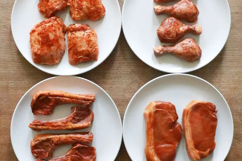 Quatre assiettes de viande crue marinée — morceaux de poulet, pilons, côtelettes et tranches d'échine — nappées d'une marinade rouge aux épices, sur une table en bois