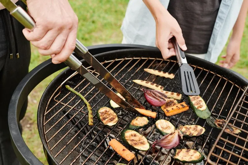 Petit barbecue rond, deux personnes retournent des rondelles de courgette, d'oignon rouge et de carotte sur la grille avec des pinces