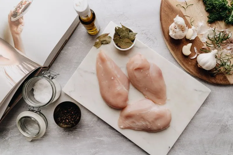 Poitrines de poulet crues sur une planche en marbre, à côté d'une planche en bois avec de l'ail et des herbes fraîches