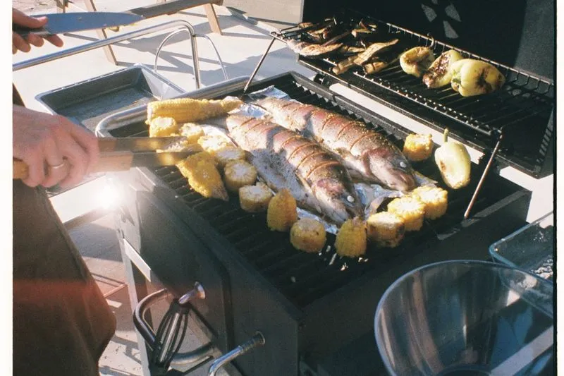 Plusieurs poissons entiers sur la grille d'un barbecue entre des épis de maïs, avec des poivrons qui grillent sur une grille voisine