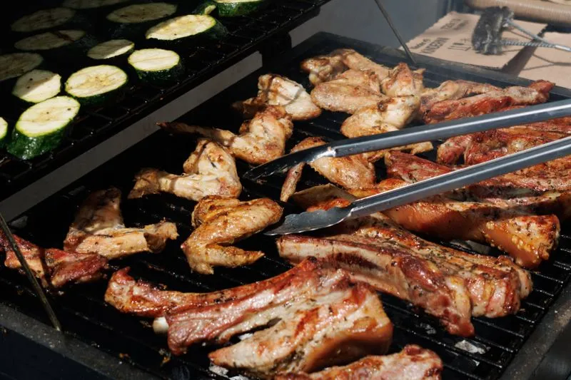 Rondelles de courgettes, ailes de poulet et travers de porc sur la grille d'un barbecue, avec une pince qui retourne la viande