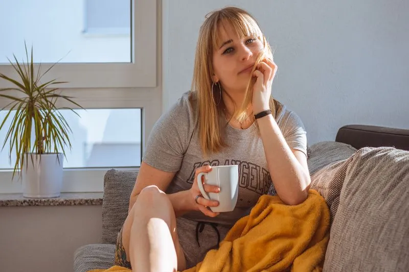 Jeune femme assise dans un canapé, un plaid moutarde sur les jambes et une tasse à la main, au soleil près d'une fenêtre