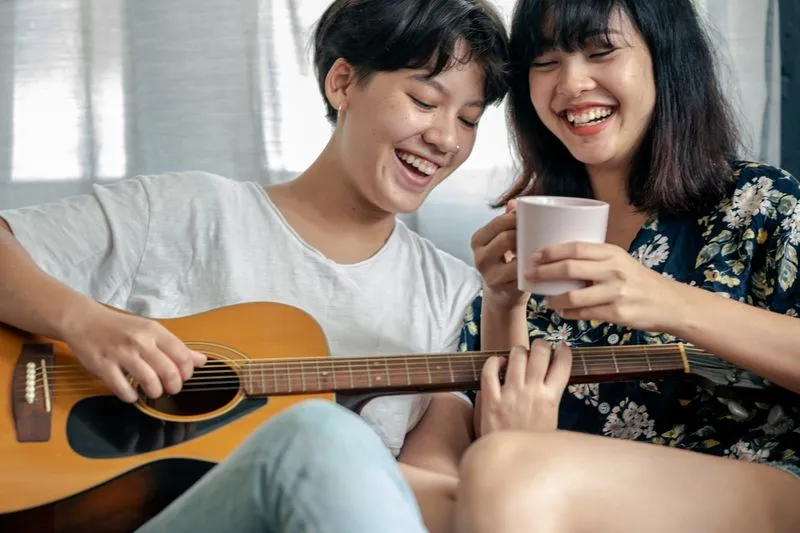 Deux jeunes femmes qui rient ensemble, l'une jouant de la guitare pendant que l'autre tient une tasse de café