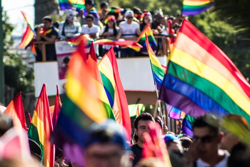 Un grand drapeau arc-en-ciel tenu à bout de bras au-dessus d'une foule, lors d'une marche des fiertés ensoleillée