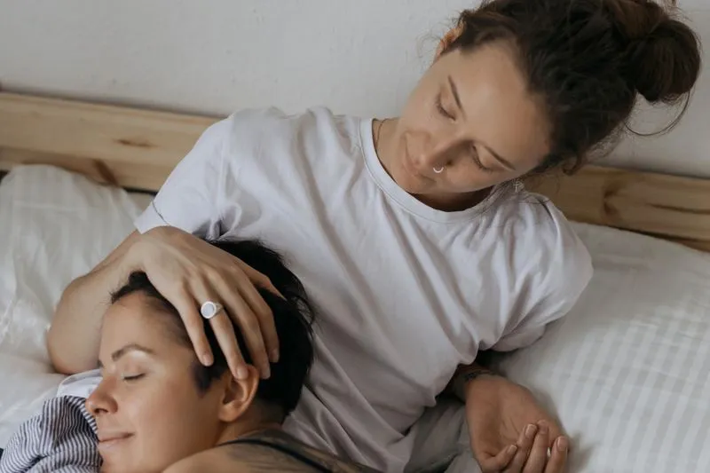 Deux femmes dans un lit aux draps clairs, l'une allongée les yeux fermés pendant que l'autre lui caresse tendrement les cheveux
