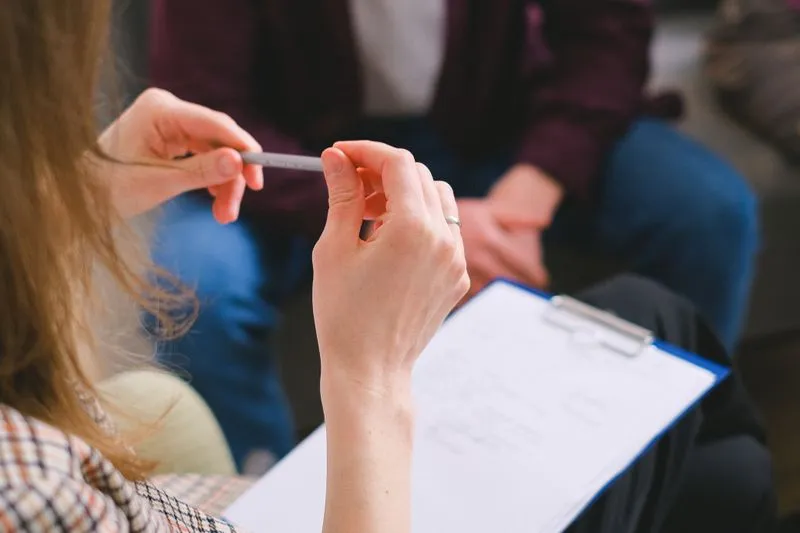 Gros plan sur les mains d'une jeune femme qui tient un stylo au-dessus d'un formulaire fixé sur une planchette, face à une personne assise en face d'elle