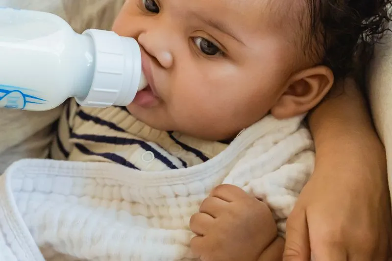 Bébé qui boit au biberon, blotti au creux du bras d'un adulte, à l'intérieur