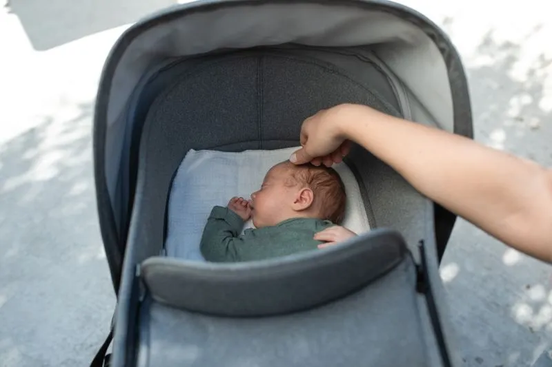 Bébé endormi dans la nacelle d'une poussette à la capote relevée, la main d'un adulte sur son front