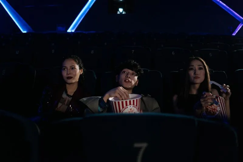 Trois amis assis dans une salle de cinéma, deux d'entre eux avec un pot de pop-corn, le visage éclairé par l'écran