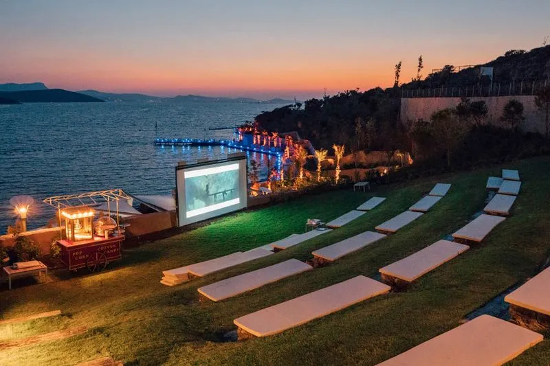 Rangées de bancs face à un grand écran de cinéma en plein air, au bord de l'eau, à la tombée de la nuit
