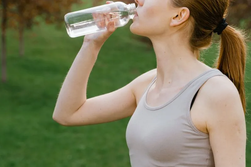 Une sportive en brassière et débardeur boit à sa bouteille d'eau dans un parc