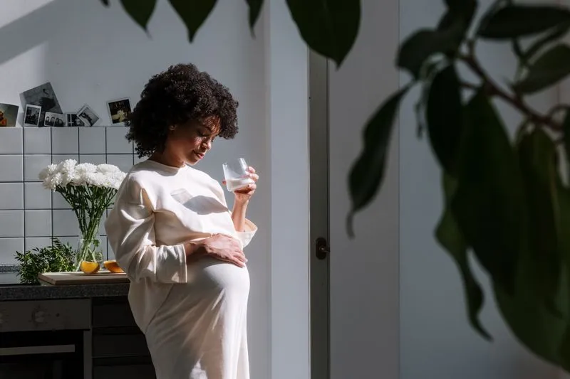 Une femme enceinte tient un verre, une main posée sur son ventre, dans une cuisine lumineuse près d'un bouquet de fleurs blanches