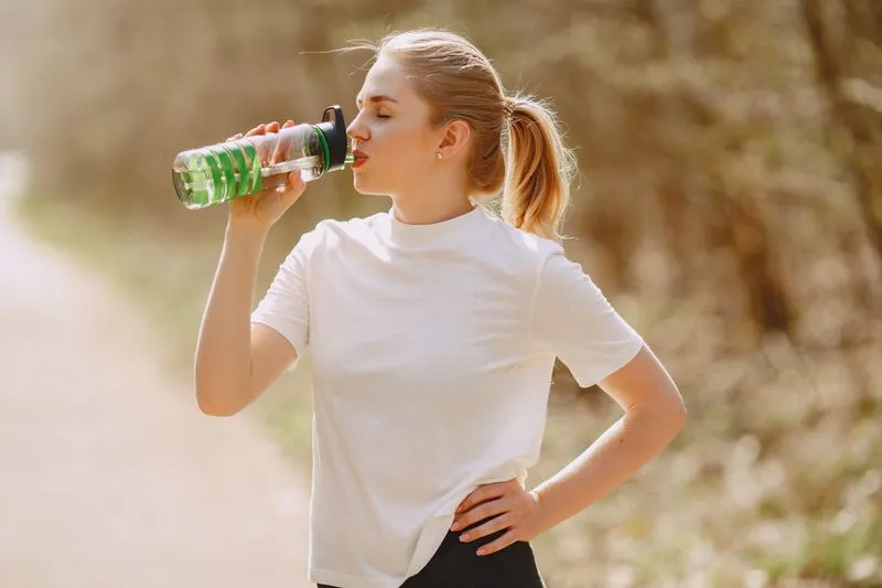 Une jeune femme en tenue de sport boit à sa gourde en extérieur, sous une lumière chaude de fin de journée