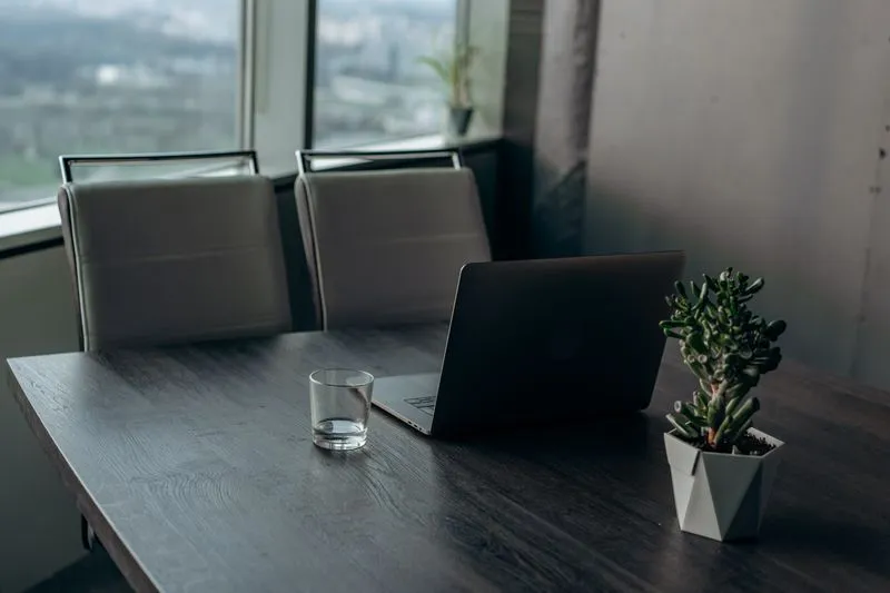 Un verre d'eau posé sur un bureau en bois sombre, à côté d'un ordinateur portable et d'une plante grasse, près d'une grande fenêtre