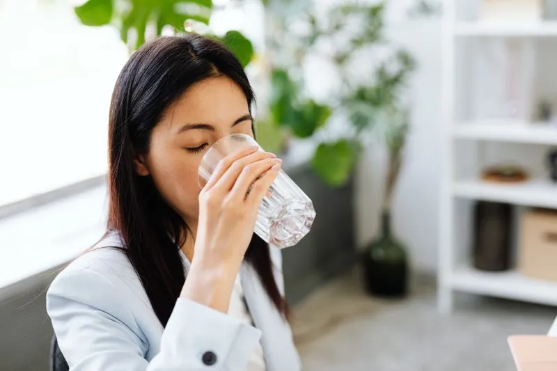 Une jeune femme boit un verre d'eau fraîche près d'une fenêtre ensoleillée