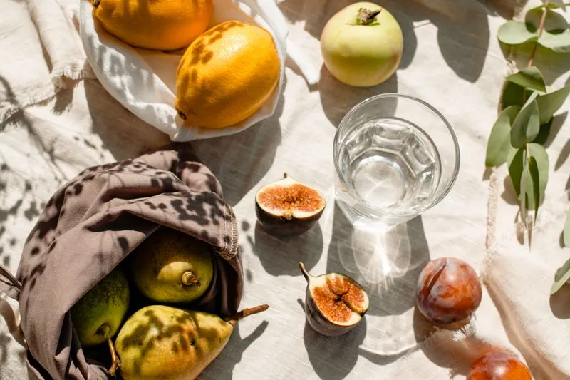 Verre d'eau entouré de fruits d'été — figues, citrons, pomme, prunes — sur un tissu de pique-nique