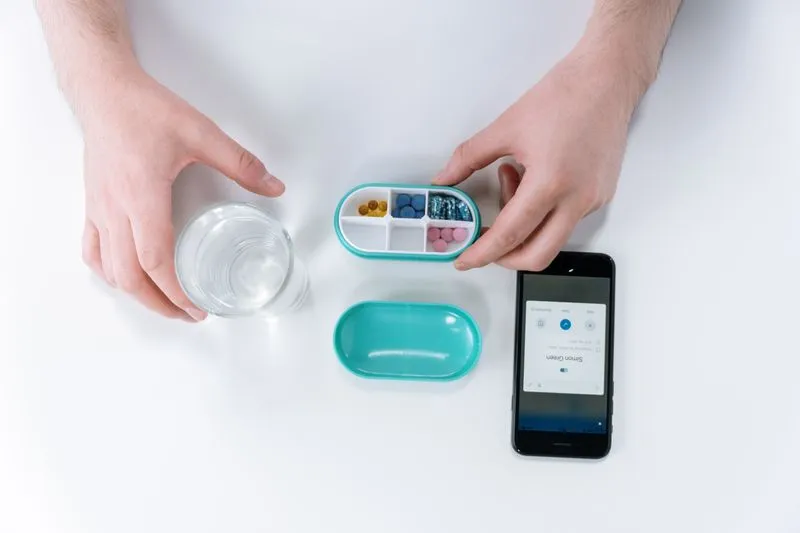 Petite boîte à pilules ouverte, tenue d'une main, à côté d'un verre d'eau et d'un smartphone
