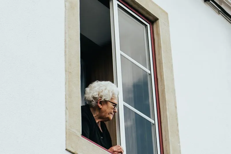 Femme âgée aux cheveux blancs et aux lunettes, accoudée à une fenêtre ouverte sur une façade blanche