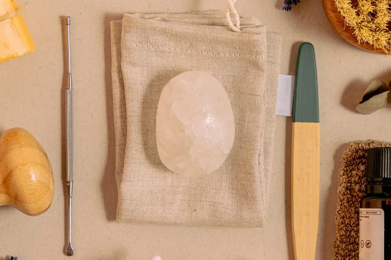 Pierre d'alun ovale et translucide posée sur un tissu en lin beige, entourée d'accessoires de soin naturels