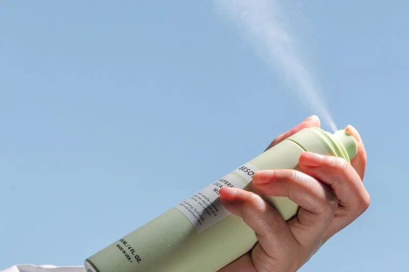 Main tenant une bombe de déodorant spray vert d'eau, qui projette un nuage fin devant un ciel bleu