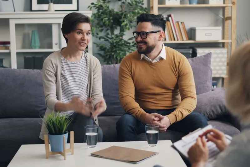 Couple assis côte à côte face à une professionnelle, dans un bureau lumineux, pendant un entretien