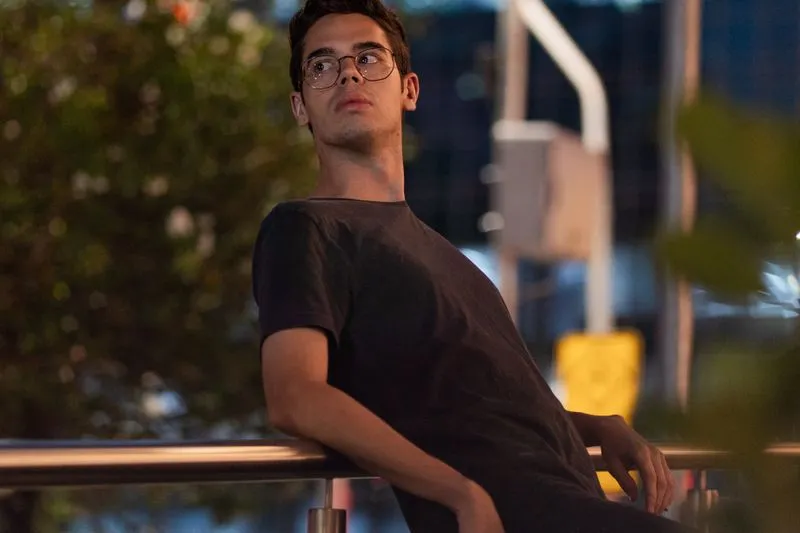 Jeune homme à lunettes accoudé à une rambarde, en ville à la tombée du jour, l'air songeur