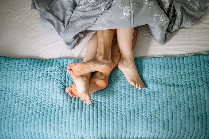 Vue de dessus de jambes de couple entremêlées sous une couette, sur un lit