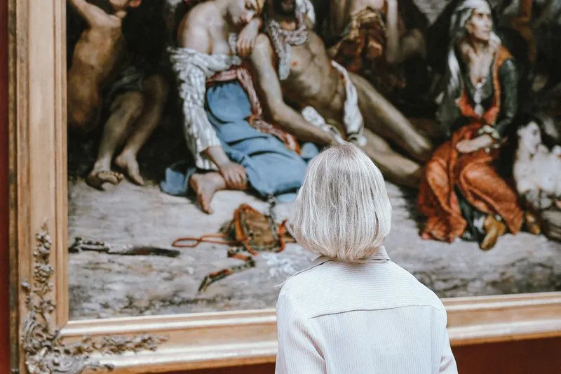 Femme aux cheveux blond platine, de dos, seule face à un grand tableau ancien dans un cadre doré