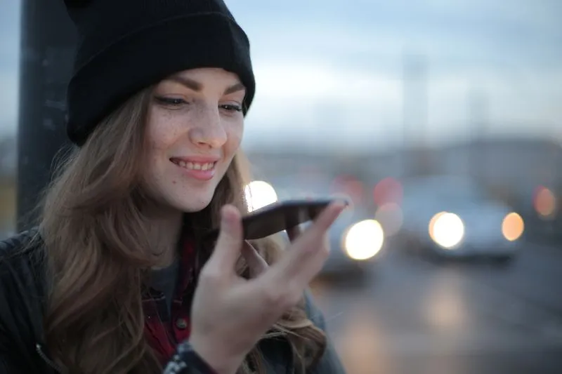 Jeune femme en bonnet noir dictant un message vocal sur son téléphone au bord d'une rue, à la tombée de la nuit, des phares flous derrière elle