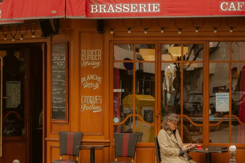 Femme en trench beige et lunettes, assise seule à la terrasse d'une brasserie, une tasse rouge devant elle et son téléphone à la main